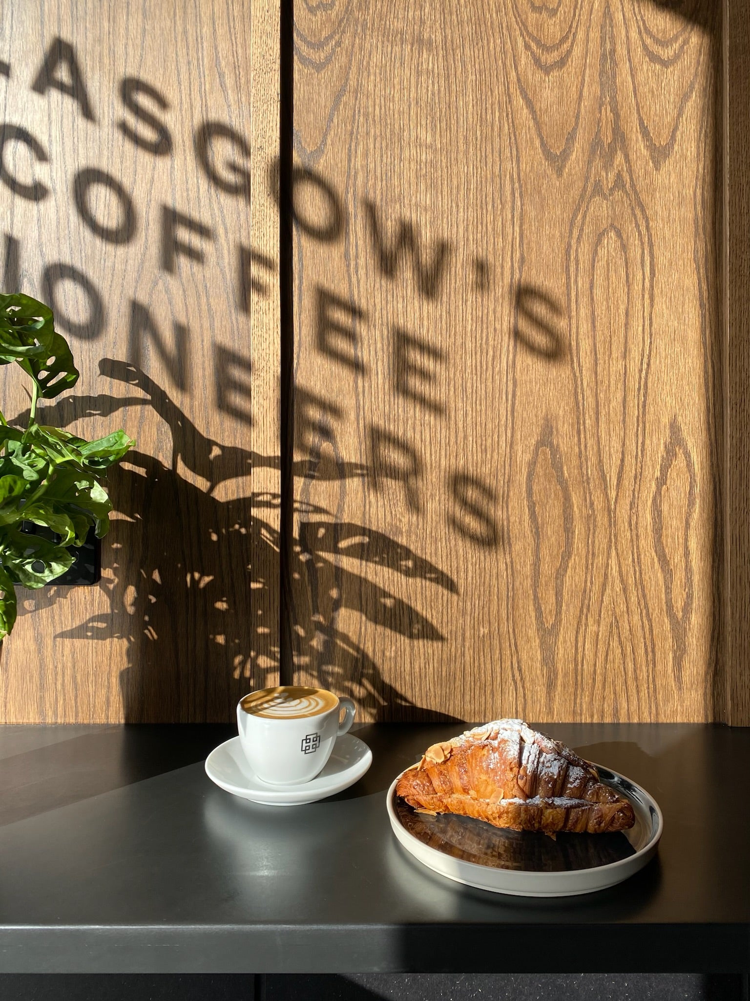 Café setting with a cup of coffee and a croissant on a table against a wooden wall with 'Glasgow's Coffee' branding.