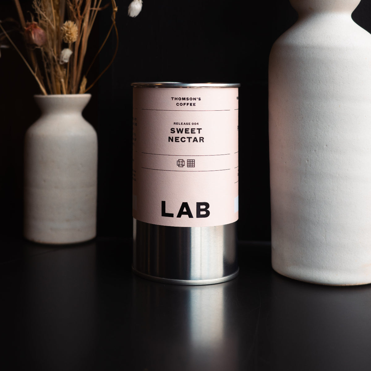 Two ceramic vases with dried flowers on a black background, featuring the LAB release 004 coffee tin labeled 'Sweet Nectar'.